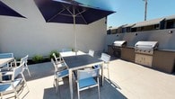 This outdoor communal space is designed for comfortable al fresco living and entertaining. The area is centered around a spacious concrete patio that accommodates multiple seating zones, creating a versatile environment for both casual gatherings and relaxed dining.

A standout feature is the built-in outdoor kitchen station, which includes a stainless steel gas grill set into a stone-topped counter with cabinet storage below — fully equipped for outdoor cooking. Adjacent to the grill, a sleek dining table with coordinating aluminum-frame chairs in blue and white is shaded by a large navy market umbrella, offering a cool and inviting spot for outdoor meals on sunny days.

Additional seating areas are arranged along the patio, including cushioned lounge chairs that provide a relaxed atmosphere for socializing. Lush ornamental grasses and mulched planting beds line the perimeter, adding greenery and softening the clean stucco walls. Wall-mounted accent lighting provides illumination for evening use.

The space opens toward a community pool area visible in the background, framed by mature trees and well-maintained landscaping. The overall design is modern and functional, with a cohesive palette of neutral tones and cool blues that create a calm, resort-like ambiance under open skies.