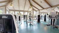 This fitness center is set within a single-story structure distinguished by a vaulted, cathedral-style ceiling with exposed wooden beams and whitewashed tongue-and-groove paneling, lending the space a clean, airy aesthetic. Ceiling fans with warm-toned light fixtures are positioned throughout, promoting comfortable airflow during workouts. Large windows line the walls on multiple sides, flooding the interior with natural light and offering views of the surrounding greenery, creating an open, welcoming environment.

The flooring consists of teal-tinted rubber tiles, providing both durability and cushioning across the workout area. A wall of mirrors along one side visually expands the space and allows users to monitor their form during exercise. The equipment selection is robust, featuring multiple Cybex International strength-training machines alongside other resistance and weight equipment, all finished in a coordinated white and black color scheme. Cardio equipment is also present.

Informational fitness charts and workout guides are posted on the walls, offering guidance to users. Decorative artwork adds a finishing touch. The combination of distinctive architecture, ample natural light, and comprehensive equipment makes this a well-rounded fitness facility.