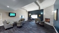 This contemporary single-floor lounge is thoughtfully designed to balance style and functionality, making it an ideal space for relaxation and social interaction. The interior is anchored by a bold deep teal accent wall, which adds a striking contrast to the neutral grey and white tones found throughout the rest of the space. Exposed cylindrical ductwork runs along the ceiling, lending an industrial character that complements the modern aesthetic.

The lounge is organized into distinct zones to accommodate a variety of activities. A cozy seating area features a mix of plush upholstered armchairs with patterned fabric, arranged around small side tables, creating an inviting spot for conversation or unwinding. Adjacent to this area, a tile-surround fireplace serves as a warm focal point, enhancing the room's ambiance. A bar-height table with matching stools provides a more casual dining or work setting, positioned near a window that allows natural light to fill the space.

A wall-mounted flat-screen television is positioned for comfortable viewing from the seating area, while a decorative chandelier and wall sconces provide layered lighting throughout. The space is finished with multi-toned dark carpeting that adds texture underfoot and ties the design elements together cohesively.