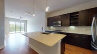 This well-appointed single-floor residence showcases a thoughtfully designed open-concept layout that seamlessly connects the kitchen, dining, and living areas. Rich, warm-toned hardwood flooring flows throughout the main living spaces, creating a cohesive and inviting atmosphere.

The kitchen is a standout feature, equipped with dark espresso cabinetry, a neutral tile backsplash, and a full suite of stainless steel appliances including a gas range, built-in microwave, and French door refrigerator. A generously sized center island with a light quartz countertop and an undermount sink serves as both a functional workspace and a natural gathering point. Stylish pendant lights hang above the island, complementing the track lighting overhead.

The living and bedroom areas are bright and airy, benefiting from large floor-to-ceiling sliding glass doors that invite abundant natural light and provide views of the surrounding greenery and cityscape. Ceiling fans in the main rooms ensure year-round comfort.

The bathroom is finished with large-format neutral tiles, a soaking tub, a separate glass-enclosed shower, and dark wood vanity cabinetry with a light countertop, providing a clean and calming retreat. Throughout the space, the neutral color palette and clean architectural lines create a modern yet welcoming environment.
