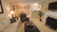This single-floor residence offers a thoughtfully designed open-concept layout that seamlessly connects the living, dining, and kitchen areas into one cohesive and functional space. The living area is anchored by plush, carpeted flooring in a warm neutral tone, complemented by a comfortable sofa, accent chair with a bold patterned upholstery, and a sleek dark coffee table that grounds the room. Soft, ambient lighting from a table lamp adds to the cozy atmosphere. The dining area sits just beyond, featuring a round table with elegant high-back chairs and a classic chandelier overhead that adds a refined touch. An ornamental wall accent adds a subtle decorative detail to the shared wall between the two areas. The kitchen is accessible via a convenient pass-through counter, offering a practical and social connection to the main living space. Light wood cabinetry and built-in appliances give the kitchen a clean, functional look. A large decorative potted plant near the window brings a refreshing natural element into the space. Throughout, the warm beige tones, carpeted floors, and soft lighting create a comfortable and welcoming ambiance ideal for everyday living and entertaining.