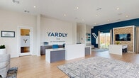Located at 4005 N Elm St in Denton, TX, the Yardly Elm by Taylor Morrison leasing and welcome center is a thoughtfully designed single-story space that serves as an inviting introduction to the community. The open-plan layout is anchored by a sleek, modern reception desk positioned beneath the prominently displayed Yardly Elm branding, set against a crisp white wall. Light hardwood flooring runs throughout, complementing the clean, contemporary aesthetic.

Bold navy accent walls create a striking visual contrast against the bright white ceilings fitted with recessed lighting, lending the space a polished, professional character. The living area features comfortable lounge seating arranged on a patterned area rug, oriented toward floor-to-ceiling sliding glass doors that flood the interior with natural light and offer direct views of the resort-style community pool and landscaped grounds beyond.

A built-in workstation with warm wood shelving, gray cabinetry, a beverage refrigerator, and under-cabinet lighting serves as a functional yet stylish amenity station. Additional windows throughout ensure natural light permeates every corner of the space. The seamless flow from reception to lounge to the outdoor-facing seating area creates a welcoming, community-oriented atmosphere that reflects the modern lifestyle Yardly Elm is designed to offer.