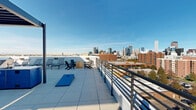 Located atop 1225 Santa Fe Drive in Denver's vibrant arts district, this expansive rooftop deck delivers a compelling outdoor experience with sweeping, unobstructed views of the Denver city skyline to the north and the Rocky Mountain range to the west. The deck is finished with large-format porcelain tiles throughout, giving the space a clean, modern aesthetic that complements its urban setting.

The rooftop is organized into several distinct functional zones. A shaded pergola structure anchors the central entertaining area, sheltering a generous dining table surrounded by teal lattice chairs — an ideal setting for al fresco meals. Adjacent to the pergola, a built-in stainless steel gas grill is housed within a bold blue outdoor kitchen station, making outdoor cooking both convenient and stylish.

An open lounge area features a comfortable sectional sofa and coffee tables, oriented to take in the panoramic mountain views. Reclining sun loungers are arranged along the perimeter railing, perfect for sunbathing beneath Colorado's famously blue skies. A cornhole board adds a recreational element to the deck, enhancing its social atmosphere.

Metal safety railings line the perimeter, ensuring open sightlines while maintaining safety. The overall design of the rooftop deck is contemporary and well-appointed, offering a versatile outdoor amenity space suited for relaxation, dining, and social gatherings.