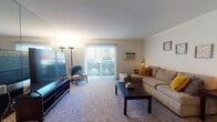 This single-floor residence offers a well-organized layout with distinct living, dining, sleeping, and kitchen areas. The living room is generously sized, featuring plush carpeting, a full-wall mirror that amplifies the sense of space, and sliding glass doors that open to a private patio, allowing natural light to fill the room. A wall-mounted air conditioning unit provides climate control. The kitchen is equipped with dark wood cabinetry, a smooth-top electric range, dishwasher, refrigerator, and a double basin sink, all set atop light wood-style vinyl flooring. Adjacent to the kitchen, the dining area features a ceiling fan with lighting and a louvered closet door for added storage. The primary bedroom is carpeted and spacious, with a large window, baseboard heating, and direct access to a private en-suite bathroom featuring a glass-enclosed walk-in shower with mosaic tile flooring and white tile walls. A second bedroom also features plush carpeting, natural light from a window, baseboard heating, and a wide louvered bi-fold closet. The second full bathroom includes a tiled tub-shower combination with mosaic tile flooring. Together, these rooms create a comfortable and functional living environment throughout.