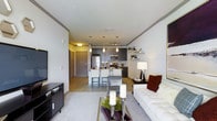 This single-floor residence showcases a thoughtfully designed open-concept layout that unifies the living, dining, and kitchen areas into one cohesive, flowing space. The living area is anchored by a plush white sofa, a glass-top coffee table, and a sleek media console, creating a comfortable yet refined atmosphere. Light hardwood flooring extends throughout, complementing the neutral, contemporary color palette of whites and cool grays.

The kitchen is a standout feature, outfitted with dark cabinetry, stainless steel appliances including a refrigerator, microwave, and range, and a kitchen island with bar-height seating for casual dining. Pendant lighting hangs above the island, adding warmth and a modern design touch. Open shelving along the wall provides both functional storage and a space for tasteful décor accents.

The space benefits from clean, smooth ceilings and well-placed recessed and accent lighting that creates a bright, welcoming ambiance throughout. The entry foyer, visible from the living area, features a paneled door and a practical layout that transitions naturally into the main living space. Overall, the residence offers a modern, urban aesthetic with a clean, organized design that balances style and everyday functionality.