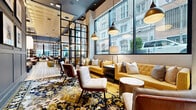 Located at 1225 Baltimore Ave in the heart of downtown Kansas City, this sophisticated hotel lobby and lounge spans a generous single floor, offering a thoughtfully curated interior that blends modern design with eclectic, artistic flair. The space is organized into distinct yet open zones connected by bold black steel-framed partitions that lend an industrial-chic character throughout.

The entry area welcomes guests with intricate patterned encaustic tile flooring, warm pendant lighting, and plush seating arrangements framed by expansive floor-to-ceiling windows that flood the space with natural light and offer dynamic views of the surrounding urban streetscape. Moving deeper into the lounge, wide-plank light hardwood floors anchor a relaxed social hub featuring a dramatic suspended conical fireplace as the central focal point, flanked by built-in bookshelves with warm accent lighting and a curated selection of books.

A dedicated games area showcases a full-size billiards table beneath a striking sputnik chandelier, surrounded by a richly patterned floral area rug in deep golds and navy. Vibrant yellow tufted sofas, cognac leather armchairs, and layered accent cushions create inviting conversation nooks throughout. Gallery-style portraiture adorns the walls, adding a bold artistic dimension. The soaring ceilings and abundance of natural light contribute to an airy, upscale ambiance that is both welcoming and visually engaging.