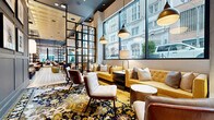 Located at 1225 Baltimore Ave in the heart of downtown Kansas City, this sophisticated hotel lobby and lounge spans a generous single floor, offering a thoughtfully curated interior that blends modern design with eclectic, artistic flair. The space is organized into distinct yet open zones connected by bold black steel-framed partitions that lend an industrial-chic character throughout.

The entry area welcomes guests with intricate patterned encaustic tile flooring, warm pendant lighting, and plush seating arrangements framed by expansive floor-to-ceiling windows that flood the space with natural light and offer dynamic views of the surrounding urban streetscape. Moving deeper into the lounge, wide-plank light hardwood floors anchor a relaxed social hub featuring a dramatic suspended conical fireplace as the central focal point, flanked by built-in bookshelves with warm accent lighting and a curated selection of books.

A dedicated games area showcases a full-size billiards table beneath a striking sputnik chandelier, surrounded by a richly patterned floral area rug in deep golds and navy. Vibrant yellow tufted sofas, cognac leather armchairs, and layered accent cushions create inviting conversation nooks throughout. Gallery-style portraiture adorns the walls, adding a bold artistic dimension. The soaring ceilings and abundance of natural light contribute to an airy, upscale ambiance that is both welcoming and visually engaging.