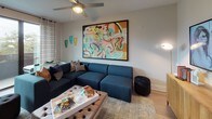 This thoughtfully designed one-bedroom, one-bathroom residence offers a cohesive blend of contemporary style and comfortable living across a single floor. The living area serves as the heart of the space, anchored by a deep navy sectional sofa and a bold, large-scale abstract artwork that infuses the room with vibrant color. Decorative wall sconces in teal and gold add artistic flair, while patterned drapes frame access to an outdoor area, allowing natural light to filter in. Light wood-look flooring runs throughout, lending warmth and continuity to the layout. The kitchen is outfitted with flat-front wood-toned cabinetry, white quartz countertops, a white tile backsplash, and a full suite of stainless steel appliances including a range, microwave, dishwasher, and refrigerator. The bedroom is a serene retreat, featuring a ceiling fan, soft neutral tones, and curated artwork that adds personality without overwhelming the calm atmosphere. The bathroom is clean and functional, with a wood-grain vanity, a large mirror with contemporary lighting, light wood flooring, and a shower-tub combination. A walk-in closet is accessible directly from the bathroom, adding practical storage to this well-rounded residence.
