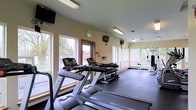 This fitness center is designed to support a well-rounded workout routine within a bright, welcoming environment. The space is lined with large windows along multiple walls, allowing abundant natural light to fill the room while offering views of the surrounding greenery outdoors — creating an energizing atmosphere for exercise.

The cardio section is anchored by a row of Matrix treadmills positioned along the windowed wall, giving users pleasant outdoor views during their workouts. A dumbbell rack provides a convenient free weights station, catering to strength training needs. Toward the rear of the space, a multi-function cable and weight machine offers options for a variety of resistance exercises, making the facility suitable for full-body training.

Overhead fluorescent lighting supplements the natural light, ensuring the space remains well-lit at any time of day. Wall-mounted televisions are positioned throughout the room, providing entertainment and keeping users engaged during their sessions. The flooring transitions to a rubberized surface, offering durability and safety underfoot. The neutral-toned walls and open layout contribute to a clean, functional, and comfortable fitness environment.