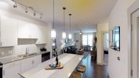 This single-floor space is designed around an open-concept layout that seamlessly connects the kitchen, dining, and living areas. The kitchen is outfitted with crisp white cabinetry, a subway tile backsplash, and sleek dark appliances, complemented by an extended white countertop that doubles as a breakfast bar with wooden stools. Three stylish pendant lights hang in a row above the counter, providing both task lighting and a design focal point, while a track lighting system along the ceiling adds further illumination to the kitchen zone.

The open layout transitions naturally into a cozy living area anchored by a navy sofa, area rug, and a ceiling fan overhead. Large windows at the far end of the space flood the room with natural light and offer views of the surrounding environment, lending an airy, welcoming atmosphere. A small dining area with a dark table and chairs is positioned near the windows, taking advantage of the natural brightness. Rich dark hardwood flooring runs throughout, providing a warm contrast to the light walls and white kitchen elements. A doorway visible to the right hints at additional rooms beyond.