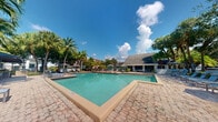Located at Lakepointe Landing in Plantation, Florida, this outdoor community amenity space showcases a generously sized swimming pool as its centerpiece. The pool features clear, blue-green water set within a well-maintained paver brick deck that surrounds the entire perimeter, offering ample space for lounging and recreation. Multiple chaise lounge chairs and seating areas are arranged around the pool, providing residents with comfortable spots to relax and enjoy the South Florida sunshine. A covered pavilion-style clubhouse structure sits adjacent to the pool area, offering shaded gathering space with open-air access to the surroundings. The space is framed by an abundance of mature tropical landscaping, including tall palm trees and lush greenery, creating a resort-like atmosphere. Umbrella-shaded seating stations are positioned throughout the deck for additional shade options. The open sky above allows for abundant natural light, complementing the vibrant, tropical character of the setting. The overall ambiance is inviting and well-suited for outdoor leisure, capturing the essence of Florida community living with a warm, relaxed atmosphere ideal for year-round enjoyment.