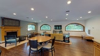 This well-appointed single-floor space is designed to accommodate a variety of activities and social gatherings. The main open area serves as the heart of the space, combining a comfortable lounge and multiple dining zones seamlessly. Distinctive arched windows with warm wood trim are a defining architectural feature throughout, framing views of surrounding greenery and filling the interior with natural light. Recessed lighting complements the natural illumination, maintaining a bright and welcoming ambiance across all areas.

The lounge section is furnished with a yellow sofa accented by colorful throw pillows, paired with light blue armchairs and a sleek console holding a wall-mounted flat-screen television. Multiple round dining tables with upholstered chairs provide ample seating for group meals or meetings. A decorative tiled fireplace with a warm wood mantel adds a focal point to one section of the main room. French doors in the dining area open to an outdoor deck surrounded by mature trees.

The full kitchen features oak cabinetry, light countertops, a central island, and stainless steel appliances. A dedicated fitness room rounds out the space, equipped with cardio machines, free weights, resistance tools, stability balls, and mirrored walls.