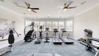 Located at 100 Monticello Mews in Hampton, Virginia, this single-floor fitness center is a comprehensively outfitted exercise facility designed to support a full range of workout routines. The space is finished with crown molding, recessed lighting, and a blue patterned carpet that lends a clean, modern aesthetic throughout.

The cardio zone is a standout feature, lined with a wide bank of white-framed windows that flood the room with natural light and offer views of the surrounding greenery. Multiple treadmills, stair climbers, and an elliptical machine are arranged to take full advantage of this bright, airy environment. Dual ceiling fans help maintain comfortable airflow during workouts.

A dedicated strength and free-weight area occupies another section of the room, equipped with a multi-station cable machine, a leg press/curl machine, adjustable weight benches, and a full rack of dumbbells and medicine balls in various weights and colors. Floor-to-ceiling mirrored walls in this zone enhance the sense of space and allow for proper form checks during exercises.

The facility also includes a water station and is accessible through glass-paneled double doors, providing a welcoming and functional entry. Overall, this fitness center delivers a well-rounded, professionally appointed space suited for residents seeking convenient, on-site exercise amenities.