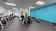 This fitness center is a thoughtfully organized, single-floor facility designed to support a comprehensive workout experience. The space is divided into distinct functional zones, each catering to different aspects of physical training. The main cardio area features a row of treadmills and elliptical machines from Matrix, positioned in front of floor-to-ceiling mirrors that visually expand the room. Wall-mounted fans provide ventilation throughout this section, enhancing comfort during intense workouts.

Adjacent to the cardio zone, a strength training area houses a selection of resistance machines, a weight bench, and a dumbbell rack, offering users a variety of options for targeted muscle training. A dedicated rowing machine occupies a quieter corner near a large window fitted with vertical blinds, allowing natural light to filter into the space.

Throughout the facility, durable rubber flooring in a dark speckled finish provides traction and impact absorption. Bright blue accent walls add visual energy to the otherwise neutral white interior, complemented by a drop ceiling with recessed fluorescent panel lighting. Strategically placed mirrors throughout the space enhance openness and allow users to monitor their form. The layout flows efficiently, with clearly marked exits ensuring safety and accessibility.
