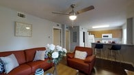 This well-appointed 2-bedroom, 2-bathroom residence offers a practical and comfortable single-floor layout designed to maximize every square foot. The living area serves as the heart of the home, featuring warm hardwood-style flooring, neutral-toned walls, and a ceiling fan with integrated lighting that keeps the space bright and well-ventilated. The room comfortably accommodates a full seating arrangement, creating an inviting space for relaxing or entertaining.

The living area flows seamlessly into the kitchen, which is outfitted with warm wood-toned cabinetry, a white refrigerator, and a built-in microwave. A raised breakfast bar with bar seating provides a casual dining option and acts as a natural divider between the kitchen and living spaces, adding both function and definition to the open layout.

Doorways visible from the living area lead to the two bedrooms and additional spaces, offering a logical and convenient floor plan. The consistent flooring throughout ties the living and kitchen areas together cohesively. With two full bathrooms serving the two bedrooms, this residence is well-suited for roommates, small families, or those who value the convenience of dual bathrooms. The overall ambiance is cozy, functional, and move-in ready.