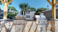Located at 2225 Ashley River Rd in Charleston, South Carolina, this outdoor amenity space is thoughtfully designed for al fresco entertaining and relaxation. The centerpiece of the area is a built-in grilling station constructed with stacked stone surround, housing dual stainless steel Coyote brand grills with ample storage cabinets below — ideal for hosting gatherings of any size. Adjacent to the grilling station, a wooden pergola provides a shaded bar area with a stone-finished countertop and vibrant orange bar seating, creating a welcoming social hub. The open-air design allows for an abundance of natural light and fresh breezes, while mature palmetto trees and lush greenery lend a distinctly Lowcountry ambiance to the space. A wrought iron fence borders the perimeter, offering both definition and openness to the surrounding landscape. Additional seating and umbrella-covered areas are visible in the background, extending the entertaining possibilities of this well-appointed outdoor retreat. The combination of durable materials, thoughtful design, and native landscaping makes this space a standout amenity reflective of Charleston's warm, outdoor-oriented lifestyle.