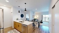 This well-appointed 3-bedroom, 3-bathroom unit in Calgary, AB offers a thoughtfully designed open-concept layout finished with warm wood-tone laminate flooring throughout. The kitchen serves as a central hub of the home, featuring a spacious waterfall-edge island with a double sink and bar seating for four, complemented by stainless steel appliances — including a full-size refrigerator, stove, and built-in microwave — as well as a combination of white upper cabinetry and natural wood lower cabinetry. Pendant lighting adds a modern, refined touch overhead. The kitchen flows seamlessly into a dining area and a comfortable living room, which is equipped with a wall-mounted mini-split climate control unit and large sliding glass doors that frame views of the surrounding neighbourhood and fill the space with natural light. Each bedroom offers a calm, neutral palette with baseboard heating and easy-care flooring. The primary bedroom benefits from a walk-through closet with open wire shelving leading directly to an ensuite bathroom. The bathrooms are clean and functional, featuring tub-shower combinations with curtained enclosures and brushed nickel fixtures. The overall design is contemporary and cohesive, balancing practicality with modern aesthetic appeal throughout.