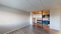 This single-floor unit offers a practical and functional layout designed for comfortable everyday living. The main living area is open and airy, featuring light gray walls, a paneled ceiling, and durable wood-look flooring that flows consistently throughout the space. A large window near the front entry allows natural light to fill the room, complemented by a wall-mounted air conditioning unit and baseboard heating for year-round comfort.

The kitchen is efficiently laid out and seamlessly accessible from the living area, separated by a partial divider with an integrated countertop that doubles as a workspace or casual dining surface. Dark navy cabinetry lines both the upper and lower sections of the kitchen, paired with a white tile backsplash and light countertops. A double stainless steel sink, an electric coil range, and a white refrigerator complete the kitchen's functional amenities.

The bedroom is a quiet, well-proportioned space with the same wood-look flooring and neutral wall tones. A bifold-door closet offers built-in shelving and hanging space for ample storage.

The bathroom is clean and straightforward, featuring a single-sink vanity with a patterned countertop, a large mirror with overhead lighting, a toilet, and a tub-shower combination. The overall design of the space prioritizes functionality and ease of maintenance.