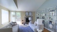 This single-floor living space offers a welcoming and well-appointed interior defined by a harmonious blend of comfort and coastal-inspired design. The living room serves as the focal point, anchored by a striking stone fireplace with a classic white mantel, above which two framed nautical prints add a subtle seaside character. Vaulted ceilings lend the room an airy, open quality, while soft blue-grey and warm beige wall tones work together to create a calm, layered atmosphere. Natural light filters generously into the space through a large sliding glass door that opens onto an outdoor deck or balcony, offering views of surrounding greenery. The living area is furnished with plush white sofas arranged around a geometric metallic coffee table, grounded by a blue area rug that reinforces the coastal aesthetic. To one side, a floor-to-ceiling glass shelving unit displays decorative objects and accessories, providing both visual interest and functional storage. Hardwood flooring runs throughout, adding warmth and continuity. Overall, the space balances relaxed elegance with practical livability.
