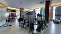 This contemporary fitness center is designed to offer a complete workout experience within a single, open-plan floor. The space is thoughtfully laid out, separating strength training and cardiovascular zones while maintaining a cohesive, open feel throughout. The strength training area features a robust selection of Precor selectorized machines, including leg press, cable systems, and multi-functional units, providing members with a full range of resistance training options. The cardio zone is visible in the background, equipped with Precor treadmills, ellipticals, and upright bikes positioned near large windows that invite generous natural light into the space. The interior design is modern and polished, with a neutral-toned rubberized floor that provides both durability and comfort underfoot. Accent walls in deep burgundy and charcoal add a bold, energizing visual contrast to the bright, airy atmosphere. Ceiling fans are installed throughout to maintain airflow, complementing the recessed lighting and cove lighting accents that lend a clean, professional ambiance. A wall-mounted television and mirrored walls enhance the functional layout, making this fitness center a well-rounded facility suited for a broad range of training needs.
