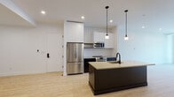 This single-floor unit showcases a thoughtfully designed open-concept layout with a contemporary aesthetic throughout. Light wood-tone flooring flows continuously across the living and kitchen areas, creating a cohesive and airy feel.

The kitchen is a standout feature, anchored by a substantial dark-finished center island with a light countertop and an integrated sink with a matte black faucet. Dual pendant lights hang above the island, adding focused illumination. The kitchen wall is finished with white grid-pattern tile backsplash, complemented by tall white upper cabinetry, dark lower cabinets, and stainless steel appliances including a French door refrigerator, built-in microwave, and range.

The living area is generously proportioned with crisp white walls, recessed lighting, and a large floor-to-ceiling window and door unit fitted with black framing, opening to a private balcony with exterior views.

The bedroom offers a clean, minimalist space with light flooring and a built-in closet equipped with shelving and hanging rods. The bathroom features grey tile flooring, white wall tile in the shower area, and black accent hardware, maintaining the unit's consistent modern design language.
