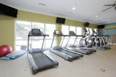 This single-level fitness center is designed to accommodate a comprehensive workout experience within a bright, organized layout. The space is immediately distinguished by its bold horizontal striped wall treatment in teal, yellow, and gray, creating an energetic atmosphere conducive to exercise. Large windows along one wall flood the interior with natural light while offering views of the surrounding outdoor area, enhancing the overall sense of openness.

The cardio section is well-appointed with multiple treadmills and elliptical trainers, each equipped with individual display screens. Wall-mounted flat-screen televisions are positioned throughout for entertainment during workouts. A multi-station cable strength machine anchors the center of the room, accompanied by a weight bench and barbell setup for resistance training. The free weight area features a fully stocked dumbbell rack alongside floor-to-ceiling mirrors that visually expand the space and aid in form monitoring.

The flooring throughout is durable, cushioned fitness-grade material suited for high-traffic use. Ceiling fans and recessed lighting provide comfortable climate control and consistent illumination. Additional fitness accessories, including a stability ball and step platforms, round out the facility's offerings.