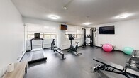 Located at 221 Queen St S in Kitchener, Ontario, this dedicated exercise room offers a fully outfitted fitness facility designed to support a comprehensive workout routine. The space features durable rubber flooring throughout, providing a safe and practical surface suited for a wide range of physical activities.

The cardio section includes a treadmill, a rowing machine, and a TRUE-brand stationary cycle, all positioned along the windowed wall. A row of windows fitted with roller blinds lines one side of the room, allowing natural light to filter in while offering views of the surrounding streetscape — a pleasant backdrop during workouts.

For strength training, the room is equipped with a TRUE functional trainer cable machine, a full dumbbell rack with a varied weight selection, and an adjustable workout bench. Stability balls add options for core and flexibility training. Wall-mounted mirrors on one side of the room enhance the sense of space and allow users to monitor their form during exercise.

Two wall-mounted flat-screen televisions provide entertainment during workouts. Flush-mounted ceiling lights ensure consistent, even illumination throughout. The crisp white walls and clean, organized layout contribute to a focused and functional training environment. Despite its compact footprint, the room is efficiently arranged to maximize usable floor space and equipment variety.