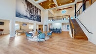 The Blakely's clubhouse is an expansive, thoughtfully designed community amenity space that immediately impresses with its dramatic double-height ceilings and warm, inviting aesthetic. The main lounge anchors the space, featuring rich hardwood flooring throughout, exposed wood beams, and a stunning coffered ceiling clad in wood planking, accented by an elegant circular chandelier. A large-scale black-and-white photographic mural of a locomotive serves as a bold artistic focal point, lending an industrial-chic character to the space. An open staircase with wood treads and metal cable railings leads to an upper mezzanine level overlooking the main floor, while floor-to-ceiling windows and French doors along one wall flood the interior with abundant natural light. A full wet bar with dark cabinetry, granite countertops, bar seating, and a stainless steel French door refrigerator provides an ideal entertaining hub. Adjacent to the main lounge, a dedicated game room offers a pool table, shuffleboard, high-top seating, and wall-mounted TVs. A separate media room is equipped with a ceiling-mounted projector, projection screen, lounge seating, and bistro tables — perfect for gatherings or movie nights. The overall ambiance blends modern farmhouse warmth with upscale community living.