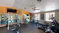 This single-floor fitness center is thoughtfully designed to support a full range of workout routines. The space is immediately distinguished by its bold orange accent walls, which energize the environment and contrast with the neutral tones throughout the rest of the room. Rubber flooring covers the entire area, providing both durability and safety during exercise.

The strength training zone is well-appointed, featuring a multi-function cable machine, a power rack with a flat bench, and a dedicated rack of dumbbells in varying weights. A large wall mirror spans one section, allowing users to monitor their form during lifts. Medicine balls, exercise balls, resistance bands, and foam rollers are neatly organized on a storage rack, offering versatile options for functional and flexibility training.

The cardio section includes multiple treadmills and elliptical machines, all positioned near large windows that invite natural light and provide views of the surrounding exterior. A wall-mounted flat-screen television adds entertainment for longer cardio sessions.

Overhead, recessed lighting and a ceiling fan ensure the space remains bright and well-ventilated. The facility is accessible via double glass doors, enhancing the flow of natural light. Framed wall art adds a finishing decorative touch, contributing to an inviting and motivating atmosphere throughout the fitness center.