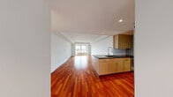 Located at 230 First Ave in Boston's Charlestown neighborhood, this one-bedroom, one-bathroom residence offers a well-designed layout across a single floor. The open-concept living and kitchen area is immediately striking, featuring rich, warm-toned hardwood flooring that runs throughout and reflects abundant natural light streaming in from large windows. The kitchen is appointed with light maple cabinetry, a dark stone countertop with a peninsula-style breakfast bar, a stainless steel refrigerator, and a modern pull-down faucet over a deep sink — all set against a tasteful tile backsplash. The spacious living area flows seamlessly from the kitchen and is framed by large windows that offer views of the surrounding Boston cityscape and what appears to be a nearby marina. The generously sized bedroom is finished with plush wall-to-wall carpeting and its own large window that continues the marina and city views, creating a bright and serene retreat. A dedicated closet provides practical storage. The bathroom features full beige tile surround in the tub/shower combination, matching tile flooring, a clean white vanity, and a frameless mirror — delivering a crisp, contemporary feel. A hallway connects the main spaces with clean transitions throughout.