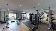 This fitness center is a thoughtfully designed, single-floor facility offering a comprehensive workout environment for all fitness levels. The space features warm wood-look flooring throughout, complemented by a neutral gray color palette and recessed lighting that creates a bright, motivating atmosphere. Large windows along the walls allow natural light to fill the room while providing views of the surrounding outdoor greenery.

The cardio section is well-appointed with Matrix-brand treadmills, elliptical trainers, stair climbers, and Spinning cycles, arranged to maximize space and usability. A dedicated strength training area includes a multi-function cable machine, a weight bench, and a dumbbell rack, all positioned adjacent to floor-to-ceiling mirrors that visually expand the space. The mirrors also serve a functional purpose, allowing users to monitor their form during workouts.

A standout design feature is the bold motivational typography mural that covers one full wall, incorporating fitness-related words and phrases in a dynamic graphic style, adding energy and visual interest to the space. The facility also includes a water station and is accessible through stylish steel-framed glass doors. Climate control is provided via ceiling-mounted HVAC units, ensuring a comfortable environment year-round. Overall, the space is clean, modern, and purposefully equipped for a complete fitness experience.