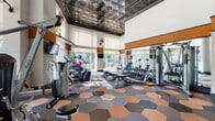 Located at 3115 Routh St in Dallas, Texas, this fitness center is a well-appointed, single-floor facility designed to support a comprehensive workout experience. The space makes an immediate impression with its soaring double-height ceilings, adorned with an ornate pressed tin ceiling detail that adds a distinctive architectural character. Bold orange accent bands run along the upper walls, complementing the crisp white surfaces and lending the space a modern, energetic aesthetic.

The floor is covered in a striking hexagonal tile pattern in tones of copper, gray, and blue, providing both visual interest and durable functionality. Floor-to-ceiling windows line the perimeter, flooding the interior with abundant natural light and offering open views of the surrounding Uptown Dallas streetscape and greenery.

The fitness floor is thoroughly equipped with a wide array of cardio machines including treadmills, ellipticals, and stationary bikes, as well as a full rack of free weights, weight benches, cable machines, and resistance training equipment. Exercise balls are also available for flexibility and core training. Wall-mounted flat-screen televisions are positioned throughout for entertainment during workouts, while industrial wall fans provide additional air circulation. A seating area near the entrance offers a comfortable transition space. The overall ambiance is bright, spacious, and motivating.