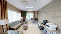 This single-floor amenity space is thoughtfully designed to serve multiple functions, blending comfort and practicality in a welcoming environment. The layout is divided into distinct zones, each catering to different needs while maintaining a cohesive aesthetic throughout.

The lounge area features a contemporary sofa and accent seating arranged around a round coffee table, creating an inviting space for relaxation or casual conversation. Nearby, a tall indoor plant and a stylish table lamp add warmth and a natural touch to the setting. Large windows dressed with neutral curtains and horizontal blinds allow generous natural light to fill the room, offering views of the greenery outside.

A high-top bistro table with sleek bar-height chairs provides an informal meeting or dining area, ideal for small group gatherings. Adjacent to this, a dedicated workstation along one wall is equipped with a desktop computer, keyboard, and printer, making it a functional resource for productivity tasks.

A distinctive reclaimed wood accent wall adds texture and visual interest, contrasting beautifully with the warm beige walls and light tile flooring. The drop ceiling with recessed panel lighting ensures consistent, even illumination throughout the space. Overall, the room strikes a balance between professional utility and comfortable, modern design.