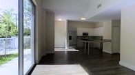 This two-story space showcases a clean, modern design characterized by an open-concept main level that seamlessly connects the living and kitchen areas. Dark hardwood flooring runs throughout, creating a rich contrast against the bright white walls and ceilings. A striking floor-to-ceiling glass wall/sliding door on one side of the living area allows an abundance of natural light to pour in, enhancing the spacious and airy feel of the interior.

The kitchen is well-appointed with dark cabinetry, granite or stone countertops, and stainless steel appliances, including a French door refrigerator with an in-door ice and water dispenser, a range, and a built-in microwave. A kitchen island or peninsula provides additional counter space and casual seating, making it a functional hub for cooking and entertaining. A tile backsplash adds texture and visual interest to the cooking area.

Recessed and flush-mount ceiling lighting ensures even illumination throughout. The overall design aesthetic is contemporary and cohesive, with a neutral color palette and high-quality finishes that contribute to a sophisticated, urban ambiance throughout the space.