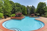 Located at 80 Newnan Lakes Blvd in Newnan, Georgia, this expansive outdoor amenity space is designed to offer a resort-like experience within a lushly landscaped setting. At the heart of the space is a generously sized, freeform swimming pool with a striking curvilinear design, bordered by elegant brick-paved decking. The pool area is thoughtfully appointed with a wide selection of lounge chairs and seating, inviting residents to relax and enjoy the outdoors. Decorative stone-accented planters and raised garden beds filled with vibrant red flowers and ornamental Japanese maples add color and texture throughout the pool surround, creating a visually dynamic environment. Mature trees frame the perimeter, providing natural shade and a sense of privacy. A covered patio adjacent to the main building offers shaded outdoor seating beneath classic columns, complemented by bistro-style tables and umbrella seating. The clubhouse building itself features a traditional Craftsman-style design with a prominent roofline, red-framed doors, and large windows. Surrounding grounds are meticulously maintained with manicured shrubs, open lawn areas enclosed by black metal fencing, and low-profile landscape lighting. The overall ambiance is serene, welcoming, and well-curated, ideal for both relaxation and social gatherings.