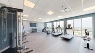 Located at 920 Manatee Ave W in Bradenton, Florida, this well-appointed fitness center offers a comprehensive exercise facility designed to meet a variety of workout needs. The single-floor space is thoughtfully laid out to maximize functionality while maintaining an open, uncluttered feel throughout.

The exercise room features a clean, modern aesthetic with soft blue-grey walls, white drop-tile ceilings with flush panel lighting, and durable grey flooring that accommodates both cardio and strength training activities. A full wall of mirrors on one side visually expands the space while aiding in form and technique during workouts.

The cardio section is equipped with multiple treadmills, elliptical trainers, a stair climber, and a rowing machine, all positioned to take advantage of the room's most distinctive feature: floor-to-ceiling windows offering elevated, panoramic views of the Bradenton cityscape and surrounding greenery. Natural light floods the space, creating an energizing atmosphere for exercise.

On the strength training side, a cable machine with a bench press attachment provides versatile resistance options, while a three-tier dumbbell rack stocked with a full range of free weights supports a wide spectrum of exercises. Wall-mounted flat-screen televisions are positioned for easy viewing from both the cardio and stretching areas. The space also includes posted facility guidelines and a clearly marked exit, ensuring a safe and organized environment.
