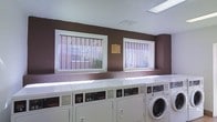 This shared laundry facility is designed with practicality and accessibility in mind. The single-floor room features a row of white commercial-grade top-loading washers and front-loading dryers lined along the wall, providing ample capacity for multiple users simultaneously. The walls are finished in a deep brown tone, giving the space a distinct, utilitarian aesthetic. Two windows fitted with decorative security grating allow natural light to filter into the room, brightening the otherwise compact interior. Vertical blinds on the windows offer adjustable light control and a degree of privacy. Overhead, a recessed fluorescent light panel ensures consistent illumination throughout the space. A posted notice board between the windows provides users with relevant rules and guidelines for use of the facility. The flooring and surfaces appear clean and well-maintained, reflecting a commitment to upkeep. Ventilation is addressed through ceiling-mounted HVAC vents, contributing to a comfortable environment during use. Overall, the laundry facility is straightforward and efficiently organized, offering residents a convenient and fully equipped space for their laundering needs.