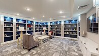 Located at 113 Ellington Blvd in Gaithersburg, MD, this elegantly designed community living room offers a comfortable and functional gathering space across a single floor. The room is anchored by floor-to-ceiling built-in bookshelves with navy blue backing and integrated accent lighting, wrapping around multiple walls to create a warm, library-inspired atmosphere. Recessed ceiling lighting and wall sconces illuminate the space evenly throughout.

The main seating area features plush upholstered sofas and armchairs arranged around a white coffee table, all set atop a patterned, textured carpet in neutral gray tones that runs throughout the room. A large flat-screen television is mounted above a row of white cabinetry along one wall, complemented by a decorative fireplace with an ornate metal screen, adding a classic focal point to the lounge area.

A separate nook accessible via glass sliding doors provides a more intimate setting, furnished with a round wooden table and gray upholstered chairs beneath a stylish chandelier — ideal for small group gatherings or casual meetings. The space flows seamlessly between its distinct functional zones, offering both relaxed lounging and social interaction in a refined, welcoming environment.