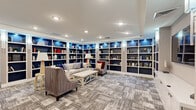 Located at 113 Ellington Blvd in Gaithersburg, MD, this elegantly designed community living room offers a comfortable and functional gathering space across a single floor. The room is anchored by floor-to-ceiling built-in bookshelves with navy blue backing and integrated accent lighting, wrapping around multiple walls to create a warm, library-inspired atmosphere. Recessed ceiling lighting and wall sconces illuminate the space evenly throughout.

The main seating area features plush upholstered sofas and armchairs arranged around a white coffee table, all set atop a patterned, textured carpet in neutral gray tones that runs throughout the room. A large flat-screen television is mounted above a row of white cabinetry along one wall, complemented by a decorative fireplace with an ornate metal screen, adding a classic focal point to the lounge area.

A separate nook accessible via glass sliding doors provides a more intimate setting, furnished with a round wooden table and gray upholstered chairs beneath a stylish chandelier — ideal for small group gatherings or casual meetings. The space flows seamlessly between its distinct functional zones, offering both relaxed lounging and social interaction in a refined, welcoming environment.