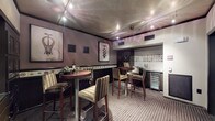 This sophisticated wine bar and tasting lounge is designed with an intimate yet refined atmosphere, making it an ideal space for entertaining and wine appreciation. The room is anchored by multiple round high-top tables paired with striped upholstered bar stools, creating a convivial setting for guests to gather and socialize.

The walls are finished in warm, muted mauve tones and adorned with framed wine-themed artwork, including striking depictions of corkscrews and wine glasses, complemented by a decorative tile border running along the lower portion of the walls. A wall-mounted wine label display adds a curated, collector's touch to the space.

Along one side of the room, dark espresso-toned cabinetry provides generous storage, with glass-fronted display cases ideal for showcasing bottles and accessories. A dedicated built-in wine refrigerator offers climate-controlled storage, while a countertop area serves as a convenient prep and serving station. Track lighting mounted to the textured ceiling highlights the artwork and casts a warm, ambient glow throughout.

The space is accessed through dark-framed glass doors from an adjacent tiled corridor, providing a clean visual transition. Patterned carpeting grounds the lounge area, adding texture and warmth underfoot. Overall, the space strikes a balance between functional wine storage and an inviting lounge environment.