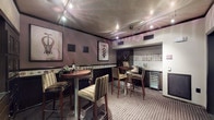 This sophisticated wine bar and tasting lounge is designed with an intimate yet refined atmosphere, making it an ideal space for entertaining and wine appreciation. The room is anchored by multiple round high-top tables paired with striped upholstered bar stools, creating a convivial setting for guests to gather and socialize.

The walls are finished in warm, muted mauve tones and adorned with framed wine-themed artwork, including striking depictions of corkscrews and wine glasses, complemented by a decorative tile border running along the lower portion of the walls. A wall-mounted wine label display adds a curated, collector's touch to the space.

Along one side of the room, dark espresso-toned cabinetry provides generous storage, with glass-fronted display cases ideal for showcasing bottles and accessories. A dedicated built-in wine refrigerator offers climate-controlled storage, while a countertop area serves as a convenient prep and serving station. Track lighting mounted to the textured ceiling highlights the artwork and casts a warm, ambient glow throughout.

The space is accessed through dark-framed glass doors from an adjacent tiled corridor, providing a clean visual transition. Patterned carpeting grounds the lounge area, adding texture and warmth underfoot. Overall, the space strikes a balance between functional wine storage and an inviting lounge environment.