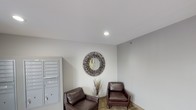This single-floor space presents a tidy and functional building lobby designed to offer residents and visitors a comfortable, organized entry experience. The lobby is finished in a clean, neutral color palette with white ceilings and light gray walls that create a bright, open atmosphere. Recessed lighting provides even illumination throughout the space.

A standout decorative element is a large, sunburst-style round mirror mounted on the wall, adding a bold artistic focal point to the room. Beneath it, a pair of rich brown leather armchairs and a slender decorative plant arrangement form an inviting seating nook, offering a comfortable spot for waiting or conversation. The flooring features a patterned tile that adds subtle texture to the overall design.

To one side, a wall-mounted bank of individual mailboxes in a brushed metal finish provides a practical and organized solution for resident mail. The space also includes a security camera, reflecting attention to building safety and resident well-being. The overall ambiance is understated yet welcoming, balancing functionality with thoughtful design details that make the lobby feel well-appointed and cared for.