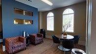 This single-story space presents a thoughtfully designed interior that balances comfort and functionality. The lounge area serves as the focal point, featuring a striking blue accent wall that contrasts with warm beige tones throughout the rest of the space, creating a visually engaging atmosphere. Decorative cutout windows are embedded within the blue wall, adding architectural interest and depth.

Natural light flows generously through two tall arched windows fitted with horizontal blinds, offering views of surrounding greenery outside. The flooring consists of patterned carpet in earthy tones, contributing warmth and texture to the room.

Seating is divided into two distinct zones. A casual lounge area includes upholstered armchairs with a rich, patterned fabric, arranged alongside a small side table — ideal for relaxed, informal gatherings. Adjacent to this is a compact dining or meeting area featuring a round table paired with modern black chairs, suitable for small group discussions or collaborative use.

A coffee or beverage station is positioned near the wall, adding a practical amenity to the space. A decorative branch arrangement serves as a subtle, tasteful centerpiece. Overall, the space offers an inviting and versatile environment.