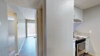 This single-story unit offers a thoughtfully arranged floor plan across two bedrooms and one bathroom. The kitchen is a functional galley-style space featuring white cabinetry with contemporary hardware, granite-look countertops, and stainless steel appliances including an electric range and refrigerator. A skylight overhead floods the kitchen with natural light, enhancing the clean, bright aesthetic. Light-toned hardwood-style flooring runs throughout, adding a cohesive and modern feel underfoot. Textured walls and a popcorn ceiling lend a classic character to the interiors. A hallway connects the main living areas, leading toward a room that benefits from natural light through vertical blinds-covered windows, offering a view of the outdoors. The overall ambiance is clean and move-in ready, with updated finishes that balance practicality and comfort. The layout makes efficient use of its footprint, providing dedicated spaces for cooking, living, and sleeping within a compact but well-organized design.