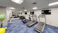 This fitness center offers a comprehensive workout environment designed to support a wide range of exercise routines. The space features a drop-panel ceiling with recessed lighting that provides bright, even illumination throughout. Distinctive blue geometric-patterned flooring ties the room together and provides a durable surface suited for active use.

The cardio zone is well-appointed with multiple treadmills, an elliptical trainer, and a spin bike, all positioned facing a row of wall-mounted flat-screen televisions — ideal for staying entertained during workouts. A Precor rowing machine adds to the variety of cardio options available.

The strength and functional training area includes a full dumbbell rack stocked with a range of free weights, a weight bench, and an organized storage rack holding medicine balls, stability balls, and resistance accessories. Large wall mirrors extend across multiple walls, visually expanding the space and allowing users to monitor their form during exercise.

The room maintains a clean, neutral aesthetic with light gray walls and a well-ventilated ceiling system. The overall layout is thoughtfully organized, ensuring ample floor space for movement between equipment. This fitness center provides a complete and functional facility for both cardio and strength training needs.
