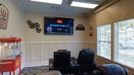 This single-floor cinema room is thoughtfully designed to deliver a comfortable and immersive entertainment experience. The space is anchored by a large wall-mounted flat-screen television, providing a clear, prominent display visible from every seat in the room. Rows of black leather recliner chairs are arranged theater-style, offering plush, supportive seating that invites guests to settle in and enjoy extended viewing sessions.

The room's interior design carries a classic cinema aesthetic, with decorative film reel wall art, a vintage-style "Cinema" sign, and themed accents that reinforce the movie-house atmosphere. The lower portion of the walls features elegant white wainscoting panels, which contrast tastefully against the warm beige upper walls, lending a polished and refined look to the space.

Natural light filters in through a window fitted with a blind, allowing for easy light control when the screen is in use. The ceiling incorporates recessed skylight panels that contribute to a bright and welcoming ambiance when the room is not in use as a screening space. Patterned carpeting completes the room, adding a layer of texture and comfort underfoot. Overall, this cinema room strikes a balance between classic movie-house charm and modern comfort.