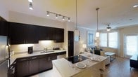 This single-floor space showcases a thoughtfully designed open-concept layout that connects the kitchen, dining, and living areas in a seamless, functional arrangement. The kitchen is a standout feature, equipped with rich dark cabinetry that contrasts elegantly against light granite countertops and a matching backsplash. A stainless steel sink, dishwasher, and black refrigerator complete the kitchen suite, while track lighting overhead ensures excellent task illumination. A large kitchen island with a built-in cooktop doubles as a breakfast bar, with pendant lights hanging above to create an inviting dining atmosphere. Place settings on the island suggest a casual yet refined dining experience. The open living area beyond is bright and airy, benefiting from natural light filtering through sizable windows dressed with blinds. A ceiling fan adds comfort to the living space, which is furnished with a sofa and accent chairs. The neutral wall tones and consistent flooring throughout the space contribute to a cohesive, modern aesthetic, making this an ideal space for both everyday living and entertaining.