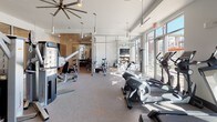 This fitness center offers a thoughtfully designed, single-level space that combines functionality with a bright, welcoming atmosphere. The main workout area is generously proportioned, featuring high ceilings with stylish pendant lighting and large ceiling fans that keep the space airy and comfortable. A bold teal accent wall lined with full-length mirrors anchors the strength training zone, which includes a variety of cable machines and free weights. The cardio section is positioned along expansive floor-to-ceiling windows, filling the room with natural light while providing open views of the surrounding area — an energizing backdrop for treadmills, ellipticals, and stationary bikes. A sleek glass partition wall elegantly separates zones within the fitness center, adding a sophisticated architectural element while maintaining visual openness throughout. The flooring transitions from plush carpet in the workout areas to warm hardwood in the adjacent children's activity room — a bright, cheerful space outfitted with a small table and colorful chairs, ideal for young ones during workout sessions. Artwork adorns the walls alongside a textured white brick accent, lending the space a modern yet inviting character. Overall, the fitness center delivers a polished, well-lit environment designed to support a full range of exercise and wellness activities.