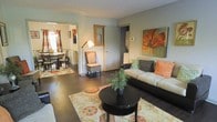 This single-floor residence offers a thoughtfully designed layout that seamlessly connects the living and dining areas, creating an open and inviting atmosphere. The living room is anchored by rich dark hardwood flooring and features a comfortable sofa accented with warm orange and sage green throw pillows, complemented by a bold floral wall canvas that reinforces the earthy, nature-inspired color palette. A round black coffee table sits atop a vibrant patterned area rug, adding texture and visual interest to the space. Natural light filters into the room, enhancing its warm and welcoming ambiance. A wicker accent chair and a standing floor lamp add layers of character and functional lighting. The dining area, visible through an open doorway, features a classic dining table and chairs set beneath a pendant light, with a large window allowing ample natural light. The neutral wall tones throughout the space provide a calm backdrop that ties the warm accent colors together cohesively. The overall design balances casual comfort with tasteful décor, making the space feel both livable and polished.