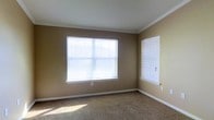 This single-story space includes a well-proportioned room finished with warm, neutral-toned walls and plush wall-to-wall carpeting in a complementary beige tone. Crown molding lines the ceiling perimeter, adding a refined architectural detail that elevates the overall finish of the space. Two windows fitted with horizontal blinds are positioned along adjacent walls, allowing generous amounts of natural light to fill the room while maintaining privacy control. White baseboards provide a clean contrast against the neutral walls, contributing to a cohesive and tidy aesthetic. The room presents a blank canvas, offering flexibility for a variety of uses such as a bedroom, home office, or sitting room. The overall ambiance is calm and understated, with the combination of soft carpeting, natural light, and classic molding details creating a comfortable and functional environment.